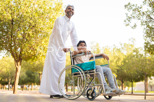 إيماءات البشاشه والمعاملة الحسنة، أب عربي سعودي خليجي يرتدي الزي التقليدي يتنزه مع ابنه المقعد، الجلوس على كرسي متحرك وقضاء الوقت في المنتزه مع العائلة، اهتمام ودعم المجتمع لذوي الاحتياجات الخاصه، تأمين احتياجات أصحاب الهمم، النظر لشيء ما، خلفية الطبيعة