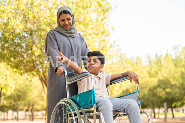 طفل مقعد يشير إل مكان ما، نمط حياة أصحاب الهمم، توفير المستلزمات الخاصة بالمعاقين، أم عربية سعودية خليجية  محجبة ترتدي عباءة رمادية تتجول في الهواء الطلق برفقة ولدها المقعد بإيماءات السعادة و السرور، الجلوس على كرسي متحرك, خلفية الطبيعة