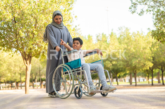 نمط حياة أصحاب الهمم، توفير المستلزمات الخاصة بالمعاقين، الاشارة إالى مكان ما، إيماءات السعادة والسرور بالخروج في الهواء الطلق، أم عربية سعودية خليجية  محجبة ترتدي عباءة رمادية تتجول في الحديقة برفقة ولدها المقعد، الجلوس على كرسي متحرك, خلفية الطبيعة