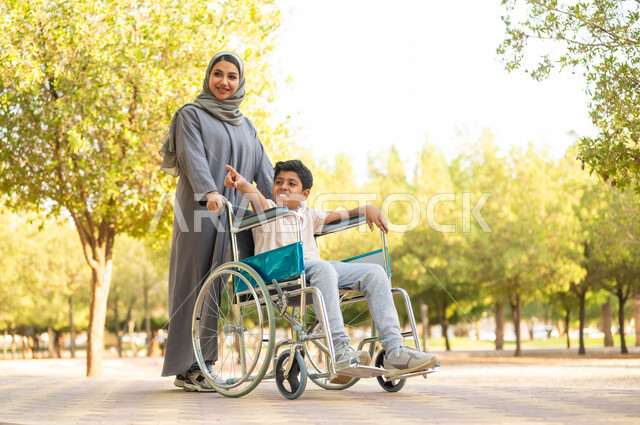 توفير المستلزمات الخاصة بالمعاقين، الاشارة إالى مكان ما، اسلوب حياة أصحاب الهمم، إيماءات السعادة والسرور بالخروج في الهواء الطلق، أم عربية سعودية خليجية  محجبة ترتدي عباءة رمادية تتجول في الحديقة برفقة ولدها المقعد، الجلوس على كرسي متحرك, خلفية الطبيعة