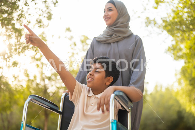 أسلوب حياة أصحاب الهمم، إيماءات السعادة والسرور بالخروج في الهواء الطلق، الاشارة إلى مكان ما، توفير المستلزمات الخاصة بالمعاقين، صورة مقربة لأم عربية سعودية خليجية  محجبة ترتدي عباءة رمادية تتجول في الحديقة برفقة ولدها المقعد، الجلوس على كرسي متحرك, خلفية الطبيعة