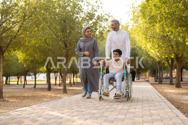 المشي في الحديقة و تبادل الأحاديث بإيماءات السرورو السعادة، الاهتمام بطفل من ذوي الهمم, قضاء لحظات ممتعة مع العائلة، الرعاية الصحية والاعتناء بالمرضى والمعاقين, نمط حياة المقعدين، أسرة عربية خليجية سعودية تتنزه في الهواء الطلق، خلفية الطبيعة