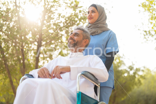 الرعاية والاهتمام بذوي الاحتياجات الخاصة، توفير معدات تساعد أصحاب الهمم على الحركة والتنقل، رجل عربي خليجي سعودي مبتسم يرتدي الثوب التقليدي يجلس على كرسي متحرك يقوم بالتجول في الحديقة بمساعدة الممرضة، جودة الحياة في السعودية، النظر الى شيء ما بايماءات السرور