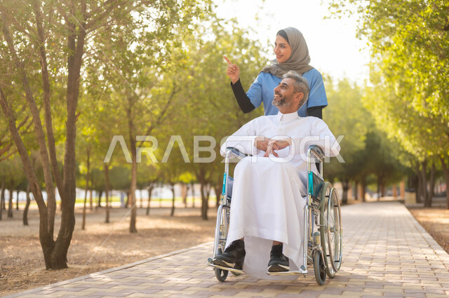 الاستمتاع بالأجواء الصيفيه المشمسة، التنزه في احدى الحدائق العامة، ممرضة عربية خليجية سعودية محجبة تقوم بمساعدة رجل مريض يجلس على الكرسي المتحرك، تأمين ادوات ومعدات تساعد أصحاب الهمم على التنقل والحركة، اسلوب الحياة في السعودية، الاشارة والنظر الى شيء ما
