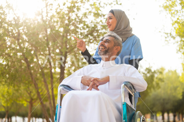 Taking a walk in one of the public parks, enjoying the sunny summer atmosphere, a veiled Saudi Arabian Gulf nurse helping a sick man sitting in a wheelchair, providing tools and equipment that help people of determination move around, the way of life in Saudi Arabia, pointing and looking at something.