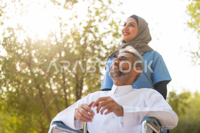 توفير ادوات تساعد أصحاب الهمم على الحركة والتنقل، صورة مقربة لرجل عربي خليجي سعودي مبتسم يرتدي الثوب التقليدي يجلس على الكرسي المتحرك يقوم بالتجول في الحديقة بمساعدة الممرضة، اسلوب الحياة في السعودية، النظر الى شيء ما، تقديم الاهتمام لذوي الاحتياجات الخاصة