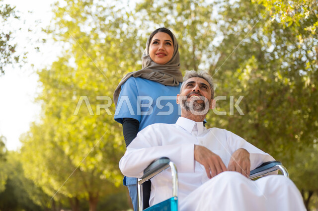 النظر الى شيء ما بتعابير السرور، تقديم الاهتمام لذوي الاحتياجات الخاصة، تأمين ادوات تساعد أصحاب الهمم على التنقل والحركة، رجل عربي خليجي سعودي مبتسم يرتدي الثوب التقليدي يجلس على الكرسي المتحرك يقوم بالتجول في الحديقة بمساعدة الممرضة، اسلوب الحياة في السعودية
