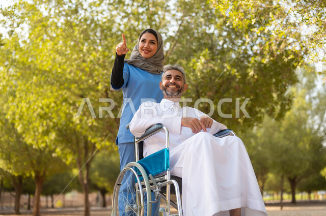 تقديم الاهتمام لذوي الاحتياجات الخاصة، تأمين ادوات تساعد أصحاب الهمم على التنقل والحركة، رجل عربي خليجي سعودي مبتسم يرتدي الثوب التقليدي يجلس على الكرسي المتحرك يقوم بالتجول والتنزه في الحديقة بمساعدة الممرضة، الاشارة الى شيء ما، اسلوب الحياة في السعودية