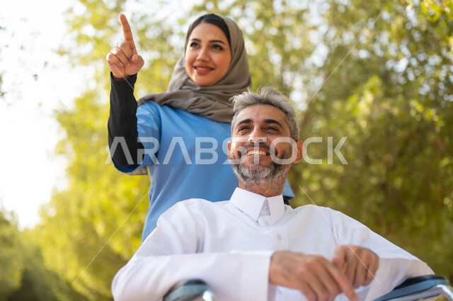Pointing to something, providing attention to people with special needs, providing tools that help people of determination move around, a close-up picture of a smiling Saudi Gulf Arab man wearing a traditional dress sitting in a wheelchair walking around in the park with the help of a nurse, lifestyle in Saudi Arabia