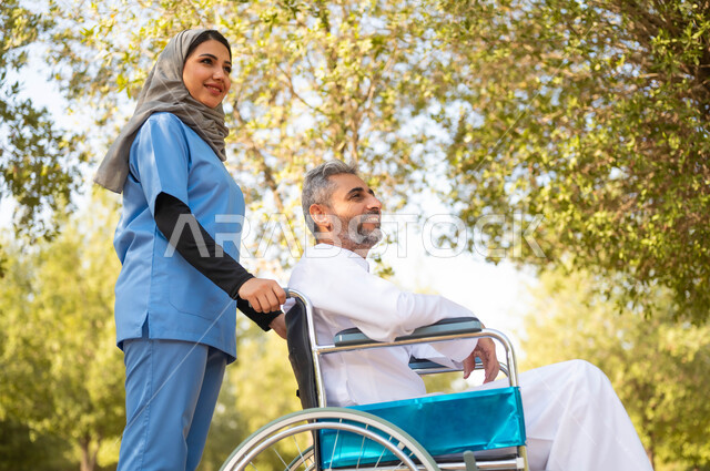 تأمين ادوات تساعد أصحاب الهمم على التنقل والحركة، الاستمتاع  بالأجواء الصيفيه المشمسة، التنزه في احدى الحدائق، صورة من الجانب لممرضة عربية خليجية سعودية محجبة تقوم بمساعدة رجل يجلس على الكرسي المتحرك، اسلوب الحياة في السعودية، الرعاية والاهتمام بذوي الاحتياجات