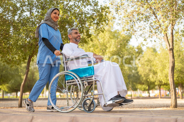 الرعاية والاهتمام بذوي الاحتياجات، تأمين ادوات تساعد أصحاب الهمم على التنقل والحركة، الاستمتاع  بالأجواء الصيفيه المشمسة، التنزه في احدى الحدائق، صورة من الجانب لممرضة عربية خليجية سعودية محجبة تقوم بمساعدة رجل يجلس على الكرسي المتحرك، اسلوب الحياة في السعودية