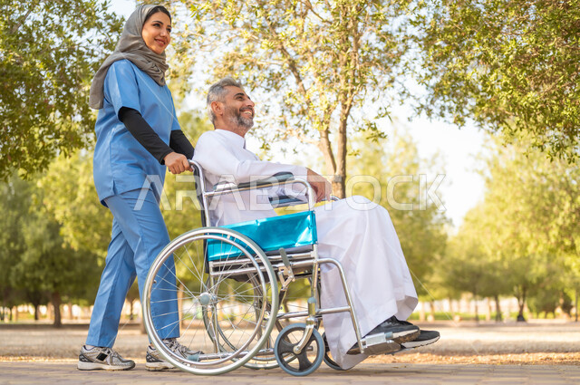 اسلوب الحياة في السعودية، الرعاية والاهتمام بذوي الاحتياجات، تأمين ادوات تساعد أصحاب الهمم على التنقل والحركة، الاستمتاع  بالأجواء الصيفيه المشمسة، التنزه في احدى الحدائق، صورة من الجانب لممرضة عربية خليجية سعودية محجبة تقوم بمساعدة رجل يجلس على الكرسي المتحرك
