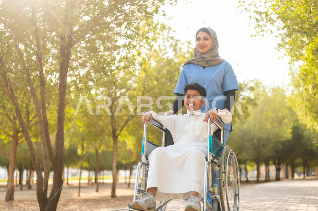 تأمين ادوات ومعدات تساعد أصحاب الهمم على التنقل والحركة، نمط الحياة في السعودية، الرعاية والاهتمام بذوي الاحتياجات الخاصة، الاستمتاع  بالأجواء الصيفيه المشمسة، التنزه في احدى الحدائق، ممرضة عربية خليجية سعودية محجبة تقوم بمساعدة ولد يجلس على الكرسي المتحرك