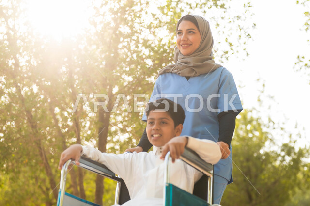الرعاية والاهتمام بذوي الاحتياجات الخاصة، الاستمتاع  بالأجواء الصيفيه المشمسة، التنزه في احدى الحدائق، ممرضة عربية خليجية سعودية محجبة تقوم بمساعدة ولد يجلس على الكرسي المتحرك، تأمين ادوات ومعدات تساعد أصحاب الهمم على التنقل والحركة، نمط الحياة في السعودية