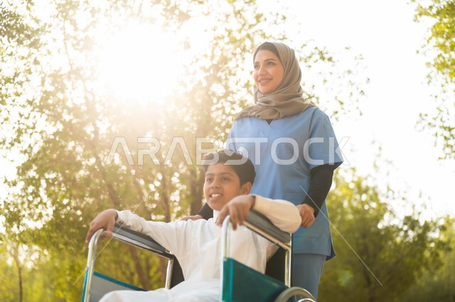 الاستمتاع  بالأجواء الصيفيه المشمسة، الرعاية والاهتمام بذوي الاحتياجات الخاصة، التنزه في احدى الحدائق، ممرضة عربية خليجية سعودية محجبة تقوم بمساعدة ولد يجلس على الكرسي المتحرك، تأمين ادوات ومعدات تساعد أصحاب الهمم على التنقل والحركة، نمط الحياة في السعودية