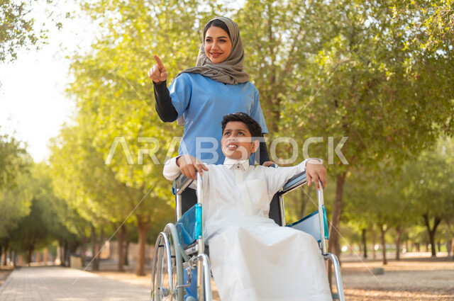 التنزه في احدى الحدائق العامة، الاشارة والنظر الى شيء ما، الاستمتاع بالأجواء الصيفيه المشمسة، ممرضة عربية خليجية سعودية محجبة تقوم بمساعدة ولد يجلس على الكرسي المتحرك، تأمين ادوات ومعدات تساعد أصحاب الهمم على التنقل والحركة، اسلوب الحياة في السعودية