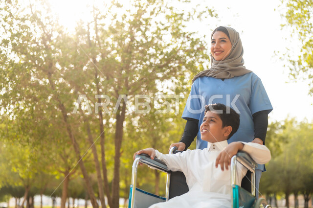 تعابير السرور لرؤية شيء ما، الاستمتاع بالأجواء الصيفيه المشمسة، ممرضة عربية خليجية سعودية محجبة تقوم بمساعدة ولد يجلس على الكرسي المتحرك، التنزه في احدى الحدائق العامة، تأمين ادوات ومعدات تساعد أصحاب الهمم على التنقل والحركة، اسلوب الحياة في السعودية