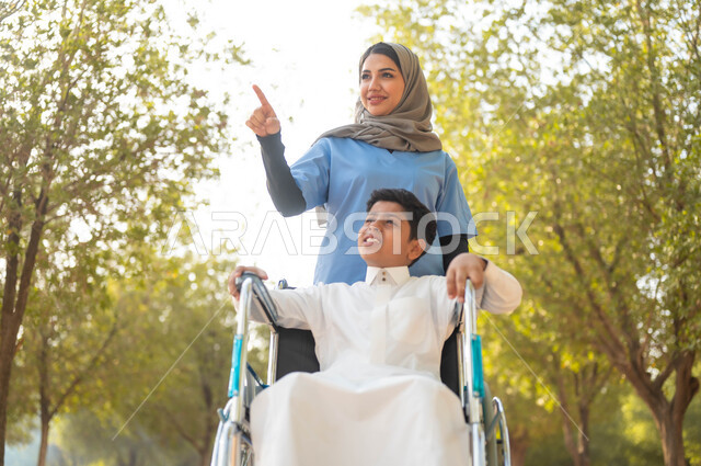 الاشارة والنظر الى شيء ما، الاستمتاع بالأجواء الصيفيه المشمسة، ممرضة عربية خليجية سعودية محجبة تقوم بمساعدة ولد يجلس على الكرسي المتحرك، التنزه في احدى الحدائق العامة، تأمين ادوات ومعدات تساعد أصحاب الهمم على التنقل والحركة، اسلوب الحياة في السعودية