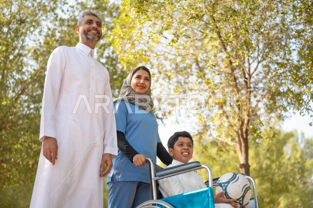Providing tools to help people of determination move around, a father enjoying a walk in a park in the summer weather with his son, getting ready to play football, taking care of people with special needs, a veiled Saudi Arabian Gulf nurse helping a boy sitting in a wheelchair, lifestyle in Saudi Arabia