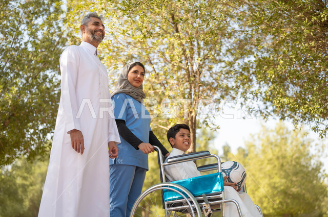 تأمين ادوات تساعد أصحاب الهمم على التنقل، أب يستمتع بالتنزه في إحدى الحدائق في الأجواء الصيفية مع ابنه، الاستعداد للعب بكرة القدم، الاهتمام بذوي الاحتياجات الخاصة، ممرضة عربية خليجية سعودية محجبة تقوم بمساعدة ولد يجلس على الكرسي المتحرك، نمط الحياة في السعودية