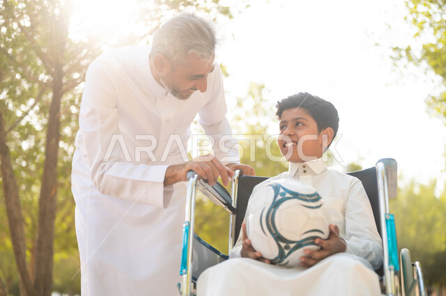 نمط حياة أصحاب الهمم، ممارسة الأنشطة الترفيهية وقضاء أوقات متعة مع الأبناء، الاستعداد للعب بكرة القدم, توفير المستلزمات الخاصة بالمعاقين، ولد عربي خليجي سعودي يرتدي الثوب التقليدي يجلس على كرسي متحرك في الحديقة برفقة والده ويقومان بتبادل الأحديث, خلفية الطبيعة.