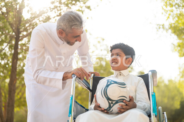 الاستعداد للعب بكرة القدم, نمط حياة أصحاب الهمم، ممارسة الأنشطة الترفيهية وقضاء أوقات متعة مع الأبناء، توفير المستلزمات الخاصة بالمعاقين، ولد عربي خليجي سعودي يرتدي الثوب التقليدي يجلس على كرسي متحرك في الحديقة برفقة والده ويقومان بتبادل الأحديث, خلفية الطبيعة.