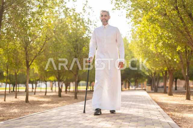 الاستجمام بالطبيعة واستنشاق الهواء الطلق، الخروج إلى أحد الحدائق العامة للترفيه عن النفس، الاستمتاع بالأجواء الصيفيه في إحدى المنتزهات، رجل شايب عربي خليجي سعودي مبتسم يرتدي الثوب التقليدي يرتكز على العكاز لسهولة التنقل، معدات تساعد على المشي.