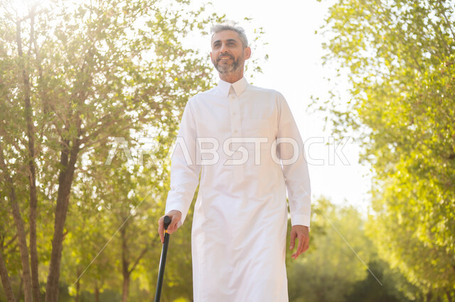 الاستجمام بالطبيعة واستنشاق الهواء الطلق، الخروج إلى أحد الحدائق العامة للترفيه عن النفس، الاستمتاع بالأجواء الصيفيه في إحدى المنتزهات، رجل شايب عربي خليجي سعودي مبتسم يرتدي الثوب التقليدي يرتكز على العكاز لسهولة التنقل، معدات تساعد على المشي.