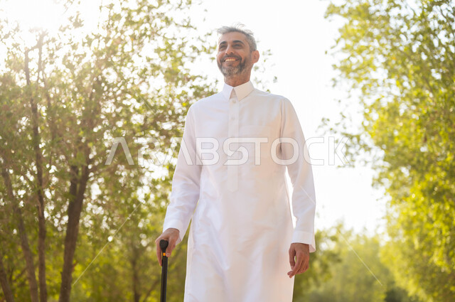 Walking around in nature and breathing the fresh air, going out to one of the public parks for recreation, enjoying the summer atmosphere in one of the parks, a smiling old Saudi Gulf Arab man wearing a traditional dress, leaning on a crutch for ease of movement, equipment that helps with walking.