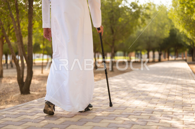 جودة حياة كبار السن في السعودية، التجول في الطبيعة واستنشاق الهواء الطلق، الاستمتاع بالأجواء الصيفيه في إحدى المنتزهات، صورة من الخلف لرجل عربي خليجي سعودي يرتدي الثوب التقليدي يرتكز على العكاز لسهولة التنقل، معدات تساعد على المشي.