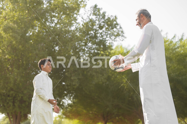 قضاء أوقات ممتعة مع الأبناء، مفهوم اللياقة والرشاقة البدنية، نمط حياة العائلة، ممارسة الأنشطة والرياضات الشبابية، أب عربي سعودي خليجي يرتدي الثوب التقليدي يقوم باللعب في الكرة مع ابنه في إحدى الحدائق العامة بإيماءات السعادة والفرح, الطبيعة الخضراء في المملكة.