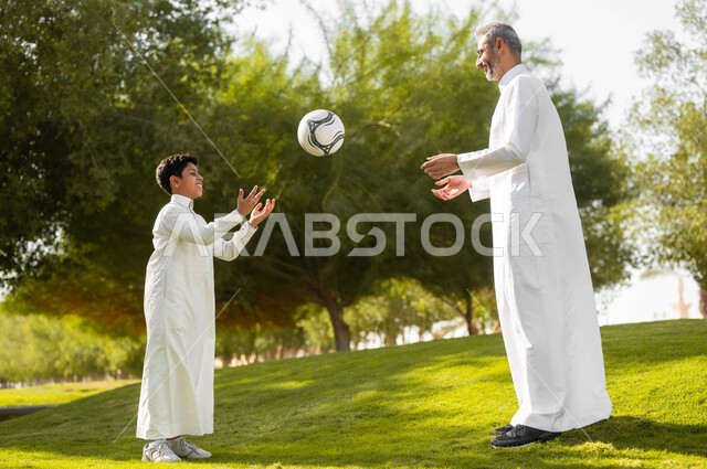 قضاء أوقات ممتعة مع الأبناء، مفهوم اللياقة والرشاقة البدنية، نمط حياة العائلة، ممارسة الأنشطة والرياضات الشبابية، أب عربي سعودي خليجي يرتدي الثوب التقليدي يقوم باللعب في الكرة مع ابنه في إحدى الحدائق العامة بإيماءات السعادة والفرح, الطبيعة الخضراء في المملكة.