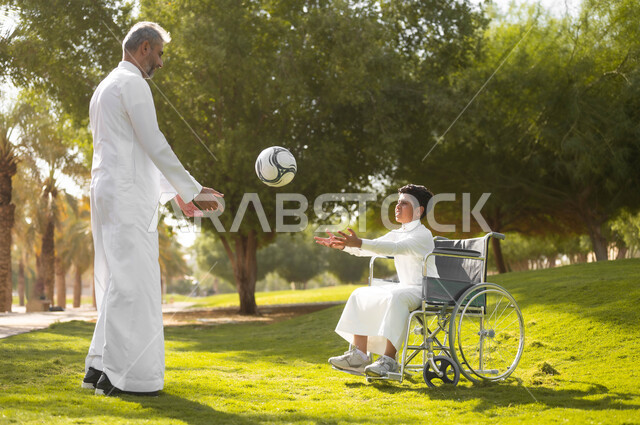 ممارسة الأنشطة الترفيهية وقضاء أوقات متعة مع الأبناء, اللعب بكرة القدم بإيماءات الفرح والسرور، توفير المستلزمات الخاصة بالمعاقين، نمط حياة ذوي الهمم، ولد عربي خليجي سعودي يرتدي الثوب التقليدي يجلس على كرسي متحرك في الحديقة برفقة والده, خلفية الطبيعة.
