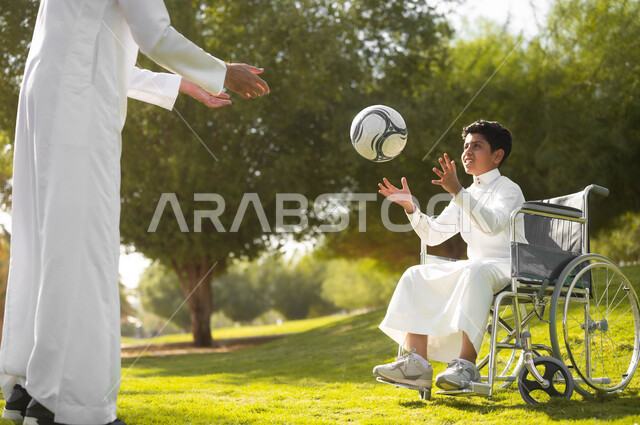 ممارسة الأنشطة الترفيهية وقضاء أوقات متعة مع الأبناء, اللعب بكرة القدم بإيماءات الفرح والسرور، توفير المستلزمات الخاصة بالمعاقين، نمط حياة ذوي الهمم، ولد عربي خليجي سعودي يرتدي الثوب التقليدي يجلس على كرسي متحرك في الحديقة برفقة والده, خلفية الطبيعة.