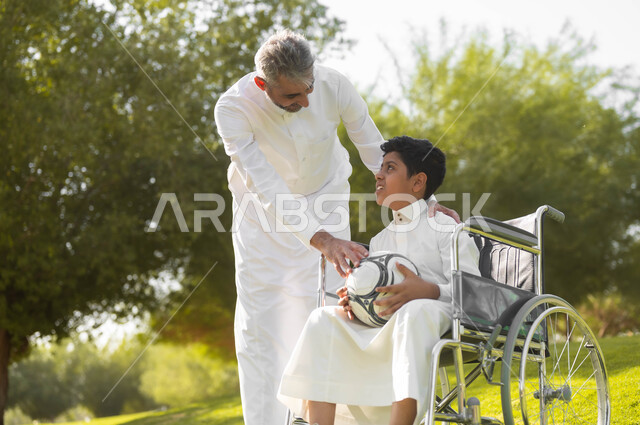 ممارسة الأنشطة الترفيهية وقضاء أوقات متعة مع الأبناء، الاستعداد للعب بكرة القدم, توفير المستلزمات الخاصة بالمعاقين، ولد عربي خليجي سعودي يرتدي الثوب التقليدي يجلس على كرسي متحرك في الحديقة برفقة والده, نمط حياة أصحاب الهمم، خلفية الطبيعة.