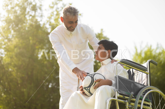 ممارسة الأنشطة الترفيهية وقضاء أوقات متعة مع الأبناء، الاستعداد للعب بكرة القدم, توفير المستلزمات الخاصة بالمعاقين، ولد عربي خليجي سعودي يرتدي الثوب التقليدي يجلس على كرسي متحرك في الحديقة برفقة والده, نمط حياة أصحاب الهمم، خلفية الطبيعة.