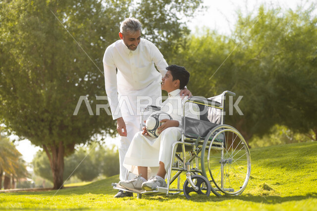 Quality of life for people of determination in Saudi Arabia, providing supplies for the disabled, preparing to play football, a Saudi Arabian Gulf father wearing a traditional dress walking around in the park with his disabled son, sitting in a wheelchair and spending an enjoyable time in the park with his family, nature background.