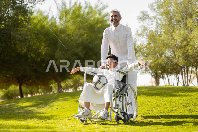 أسلوب حياة أصحاب الهمم، إيماءات السعادة والسرور بالخروج في الهواء الطلق، الاستعداد للعب بكرة القدم, توفير المستلزمات الخاصة بالمعاقين، أب عربي سعودي خليجي يرتدي الثوب التقليدي يتجول في الحديقة برفقة ولده المقعد، الجلوس على كرسي متحرك, خلفية الطبيعة.