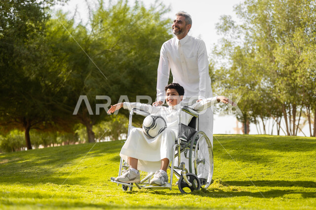 ممارسة الأنشطة الترفيهية وقضاء أوقات متعة مع الأبناء، الاستعداد للعب بكرة القدم, توفير المستلزمات الخاصة بالمعاقين، ولد عربي خليجي سعودي يرتدي الثوب التقليدي يجلس على كرسي متحرك في الحديقة برفقة والده, نمط حياة أصحاب الهمم، خلفية الطبيعة.