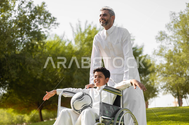 أسلوب حياة أصحاب الهمم، إيماءات السعادة والسرور بالخروج في الهواء الطلق، الاستعداد للعب بكرة القدم, توفير المستلزمات الخاصة بالمعاقين، أب عربي سعودي خليجي يرتدي الثوب التقليدي يتجول في الحديقة برفقة ولده المقعد، الجلوس على كرسي متحرك, خلفية الطبيعة.