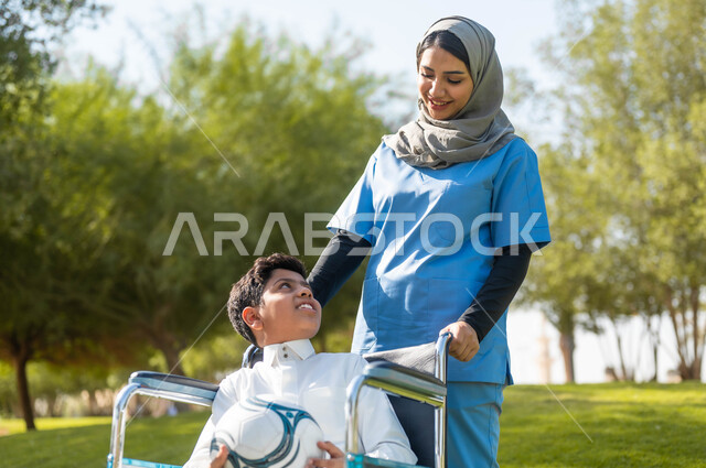 التنزه في احدى الحدائق، الاستعداد للعب بالكرة، الاهتمام بذوي الاحتياجات الخاصة، الاستمتاع  بالأجواء الصيفيه المشمسة، ممرضة عربية خليجية سعودية محجبة تقوم بمساعدة ولد يجلس على الكرسي المتحرك، تأمين ادوات تساعد أصحاب الهمم على التنقل، نمط الحياة في السعودية