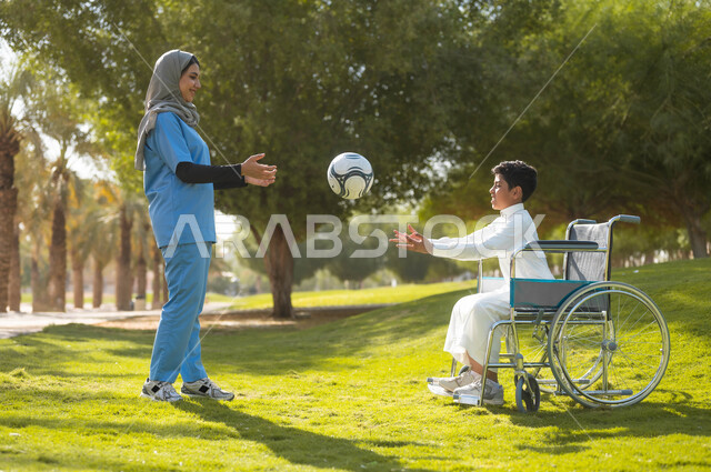 تأمين أدوات ومعدات تساعد أصحاب الهمم على التنقل والحركة، نمط الحياة في السعودية، الرعاية والاهتمام بذوي الاحتياجات الخاصة، الاستمتاع  بالأجواء الصيفيه المشمسة، ممرضة عربية خليجية سعودية محجبة تقوم باللعب بالكرة مع ولد يجلس على الكرسي المتحرك في الحديقة
