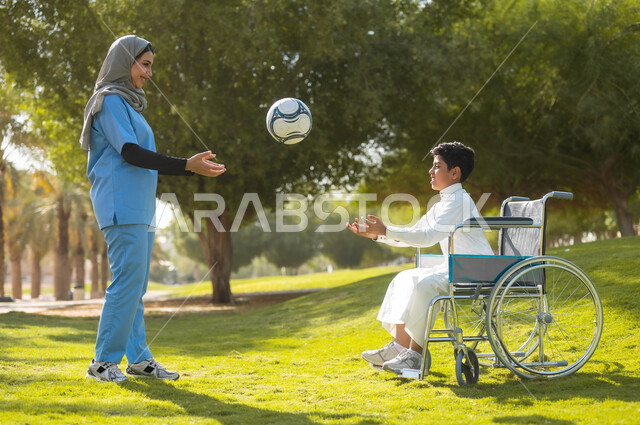 الاستمتاع  بالأجواء الصيفيه المشمسة، تأمين أدوات ومعدات تساعد أصحاب الهمم على التنقل والحركة، نمط الحياة في السعودية، الرعاية والاهتمام بذوي الاحتياجات الخاصة، ممرضة عربية خليجية سعودية محجبة تقوم باللعب بالكرة مع ولد يجلس على الكرسي المتحرك في الحديقة