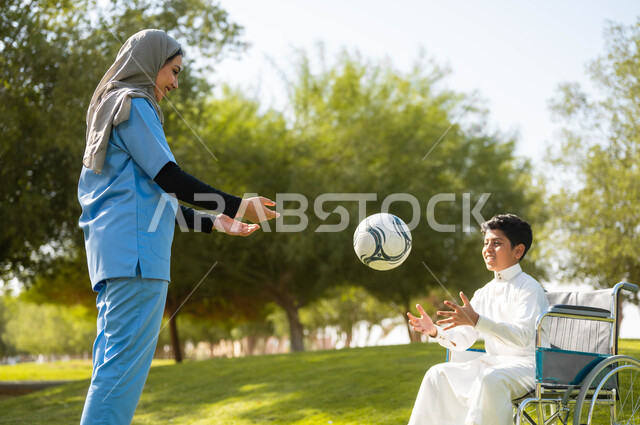 تأمين أدوات ومعدات تساعد أصحاب الهمم على التنقل والحركة، نمط الحياة في السعودية، الرعاية والاهتمام بذوي الاحتياجات الخاصة، الاستمتاع  بالأجواء الصيفيه المشمسة، ممرضة عربية خليجية سعودية محجبة تقوم باللعب بالكرة مع ولد يجلس على الكرسي المتحرك في الحديقة