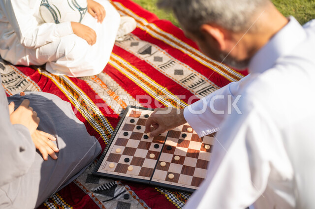 الجلوس في الطبيعة الخضراء، مفهوم التحدي والتركيز، عائلة عربية خليجية سعودية تقضي أوقات ممتعة في إحدى الحدائق في السعودية، أنشطة عائلية في الأجواء الصيفية، نزهه في الهواء الطلق، صورة مقربة لرجل عربي خليجي سعودي يلعب لعبة الضامة