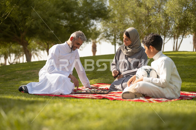 مفهوم التحدي والتركيز، عائلة عربية خليجية سعودية تقضي أوقات ممتعة في إحدى الحدائق في السعودية، أنشطة عائلية في الأجواء الصيفية، نزهه في الهواء الطلق، زوجان عربيان خليجيان سعوديان يلعبان لعبة الضامة، الجلوس في الطبيعة الخضراء