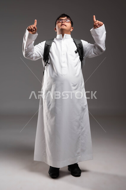 الاشارة بأصابع السبابة الى شيء ما، بورتريه لشاب عربي خليجي سعودي مصاب بمتلازمة داون يرتدي الثوب التقليدي والنظارات الطبية و يحمل الحقيبة المدرسية و ينظر الى الاعلى، دمج ذوي الاحتياجات الخاصة في مدارس السعودية، صورة لكامل الجسم، خلفية رمادية .