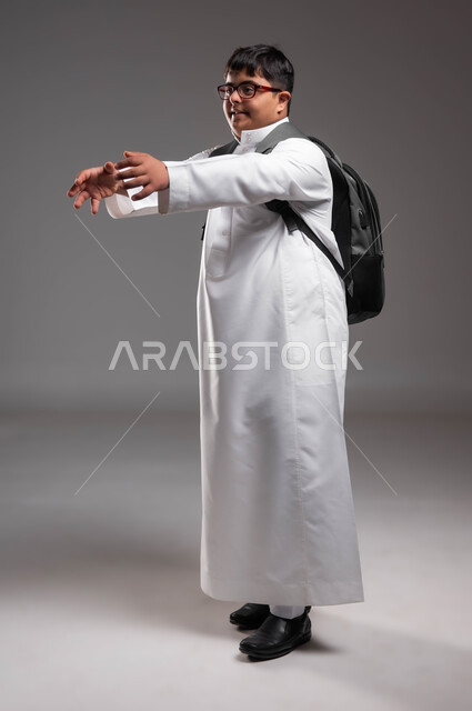 الفرح عند رؤية شخص ما والرغبة بالمعانقة، الاهتمام بذوي الهمم لشعورهم بالانتماء، بورتريه لشاب عربي خليجي سعودي مصاب بمتلازمة داون يرتدي الثوب التقليدي والنظارات الطبية و يحمل حقيبة الظهر المدرسية يمد يديه للأمام بإيماءات السعادة، صورة لكامل الجسم، خلفية رمادية .