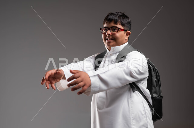 المحاولة لامساك شيء ما، بورتريه لشاب عربي خليجي سعودي مصاب بمتلازمة داون يرتدي الثوب التقليدي والنظارات الطبية و يحمل حقيبة الظهر المدرسية يمد يديه للأمام بإيماءات الفرح عند رؤية شخص ما للمعانقة، الاهتمام بذوي الهمم لشعورهم بالتقدير، خلفية رمادية .