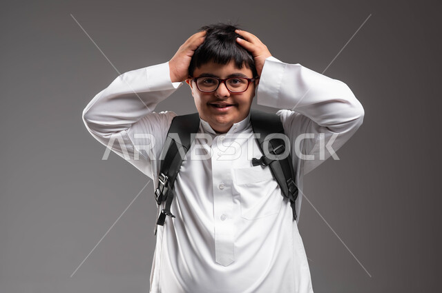 رفع اليدين للرأس بايماءات الاندهاش، بورتريه مقرب لشاب عربي خليجي سعودي مصاب بمتلازمة داون يرتدي الثوب التقليدي والنظارات الطبية و يحمل الحقيبة المدرسية وينظر الى الكاميرا، دمج ذوي الاحتياجات الخاصة في مدارس السعودية، تطور وتقدم المناهج التعليمية، خلفية رمادية .